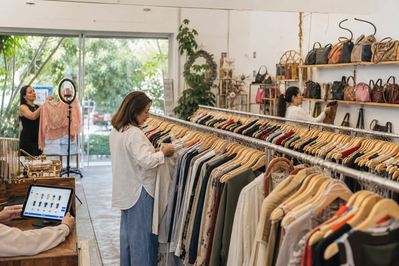 Tienda de consignación amplia y luminosa con percheros curados, exhibidores de bolsas y un rincón de transmisión